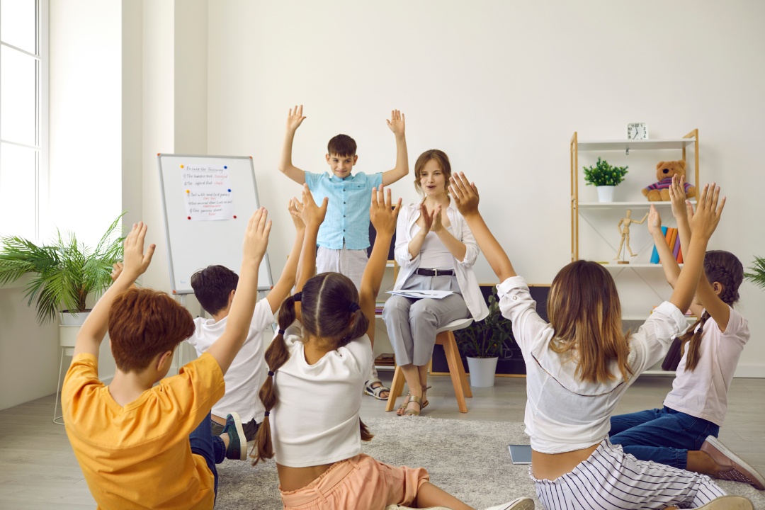 Students and teacher having fun in class. Kids playing charades or Simon Says game at school. Small group of children sitting on floor in classroom, looking at classmate and repeating his movements