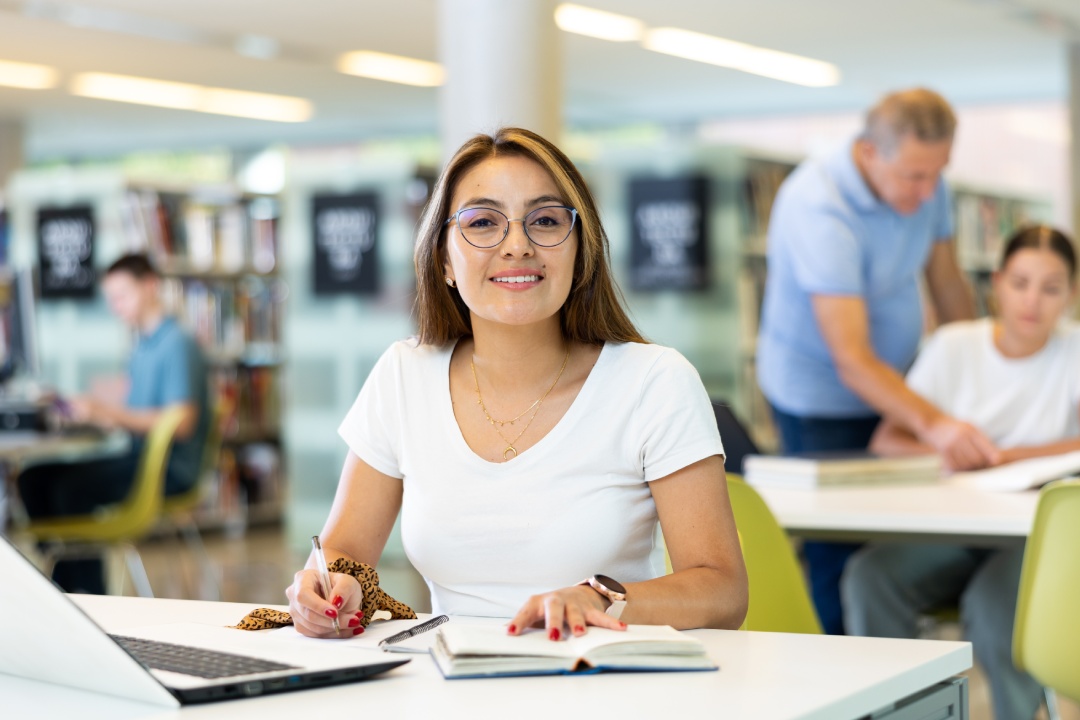 Portrait of adult latino woman studying at library using books and laptop computer