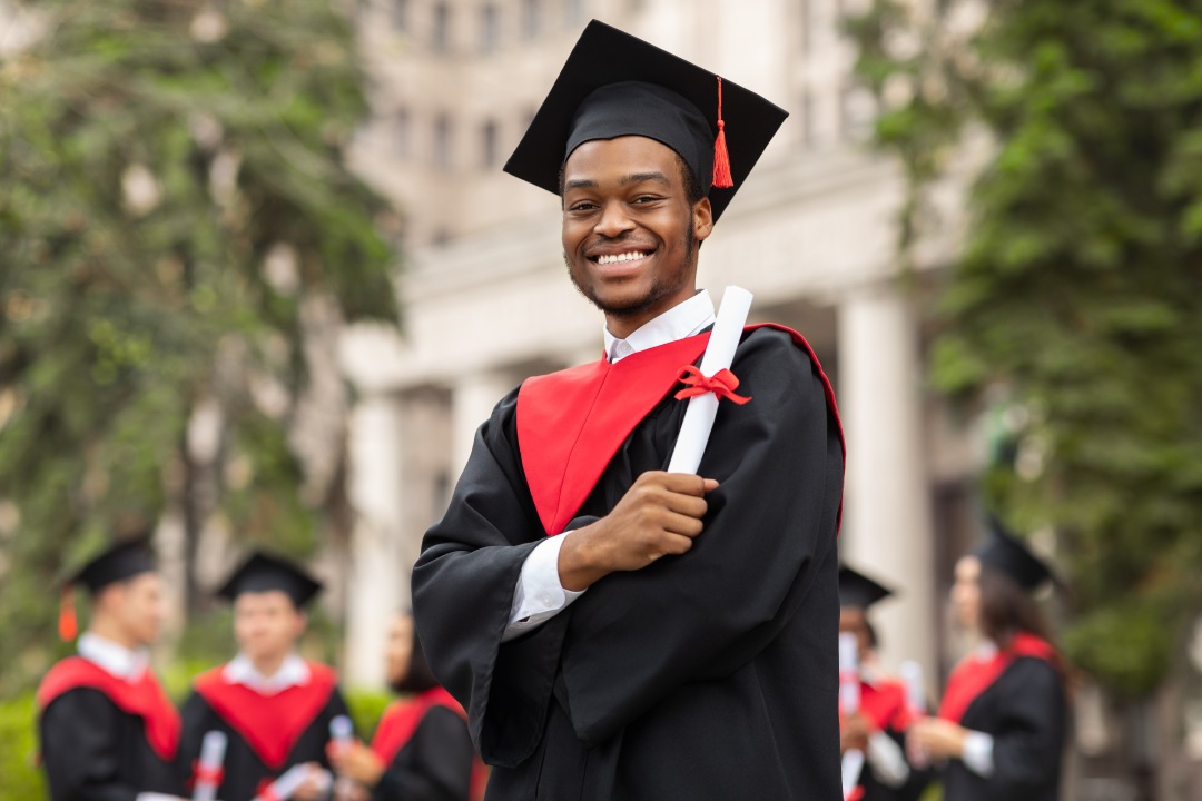 Cheerful african american guy in graduation costume showing his diploma and smiling at camera, black male student posing over international group of students at university campus, copy space