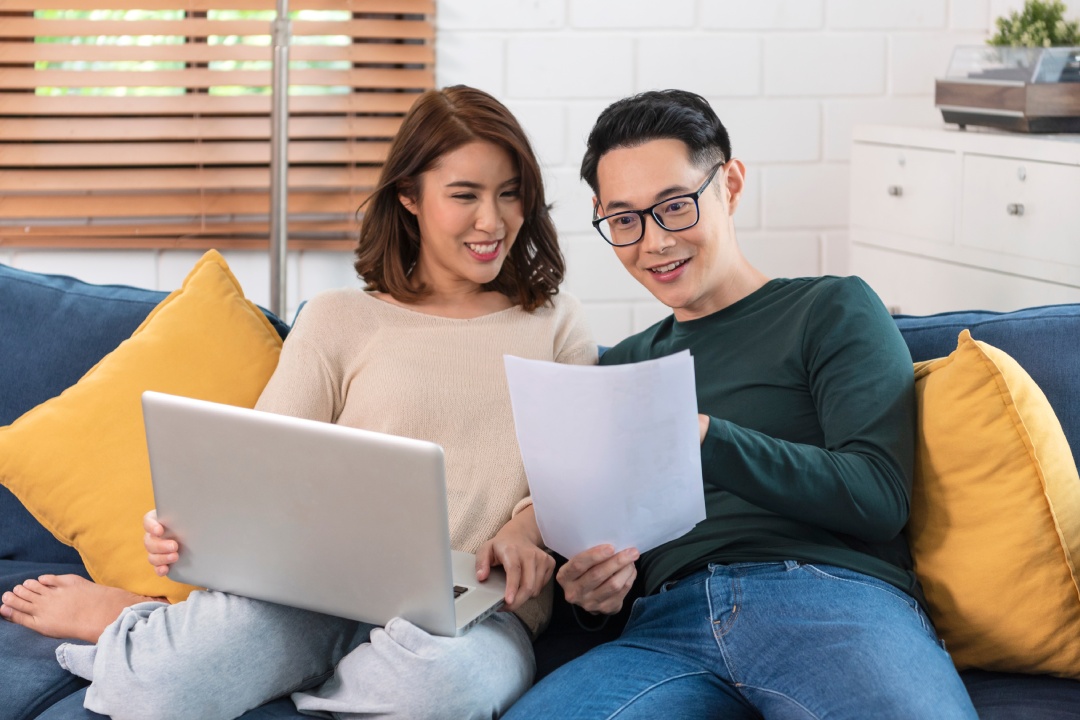 Happy Asian husband checking analyzing statement utilities bills sitting together at home.