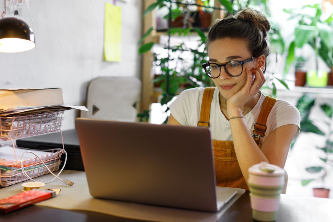 Joven jardinera con gafas que utiliza un ordenador portátil, se comunica por Internet con un cliente en el jardín de su casa/invernadero, con una taza de café/té reutilizable sobre la mesa.