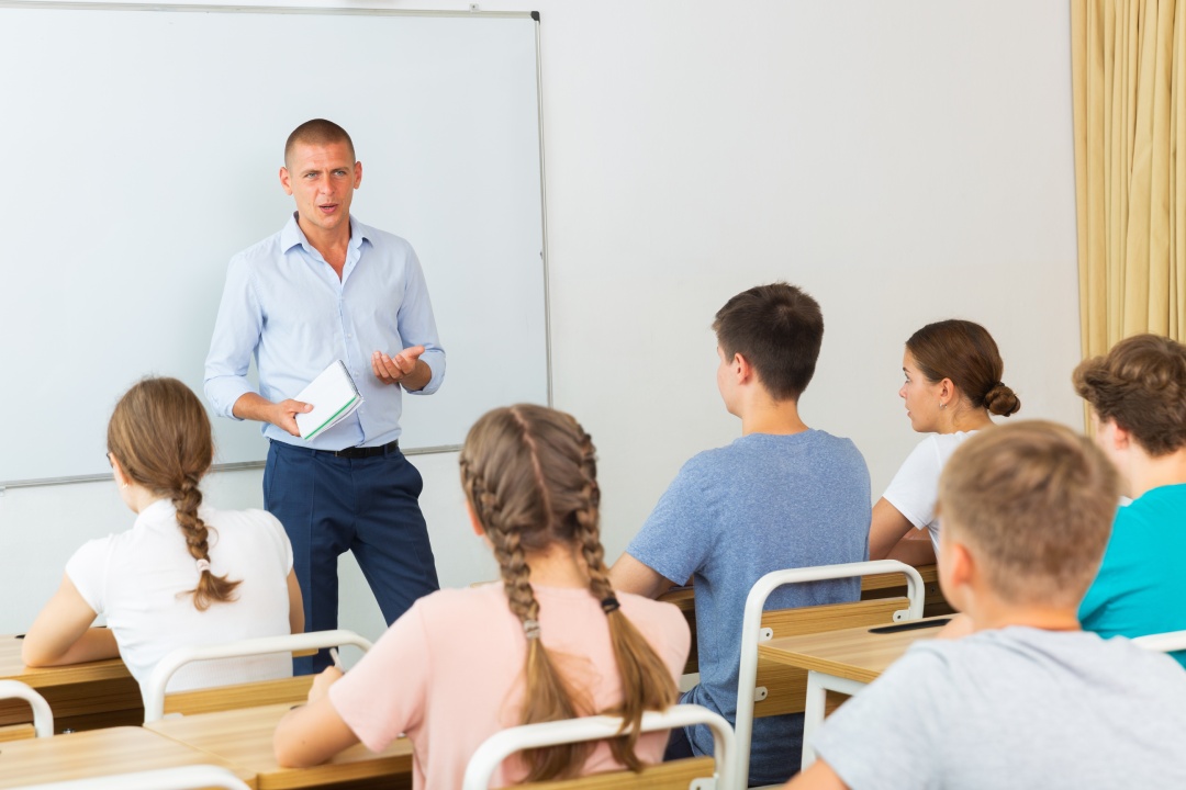 Teacher explaining studying material to teenagers during lesson at school, standing at whiteboard..