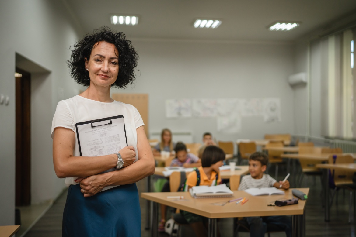 Portrait of mature teacher stand and hold clipboard with students in the background