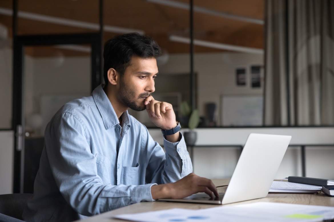 Joven directivo indio, pensativo, trabajando con un portátil, sentado en la mesa del trabajo, mirando la pantalla, tecleando, tocándose la barbilla, pensando en contenidos online, utilizando el ordenador para comunicarse en el trabajo.