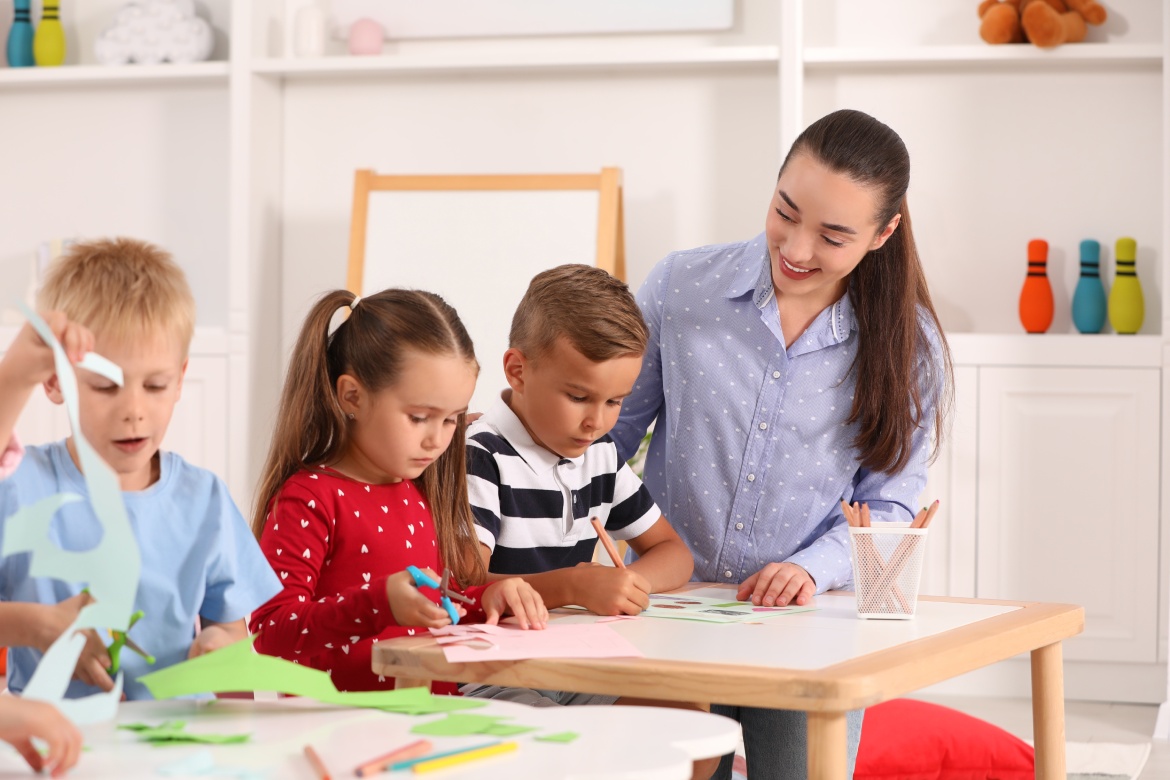 Nursery teacher and group of cute little children playing at desks in kindergarten. Playtime activities