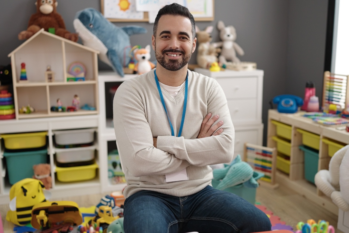 Young hispanic man teacher smiling confident sitting with arms crossed gesture at kindergarten