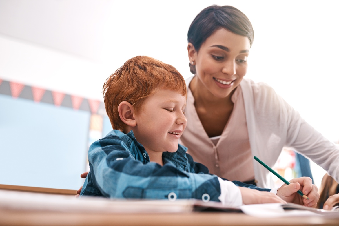 Writing, teacher and child in classroom for help in student growth, child development and advice for education. Learning, woman and boy at desk with exam notes, support and study at elementary school