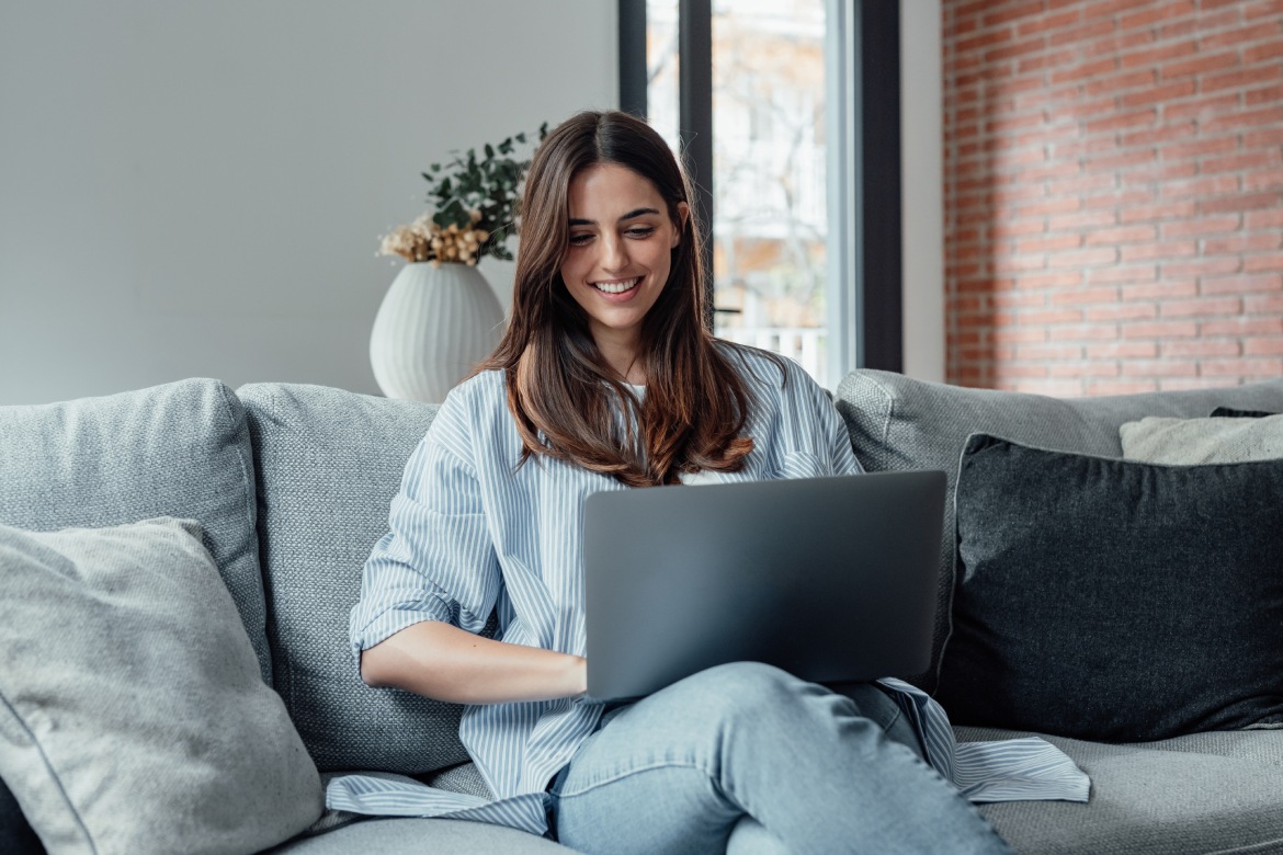 Concentrated millennial girl sit on couch working on laptop browsing internet at home during lazy weekend, focused young woman freelancer busy using computer surfing wireless web shopping online