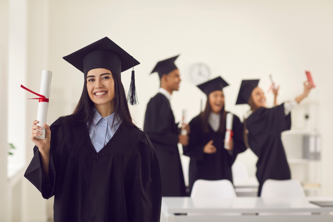 Female graduate dressed in a bachelor's gown shows the camera a diploma in her hands while standing in the classroom against the background of classmates who is taking a selfie. Education concept.