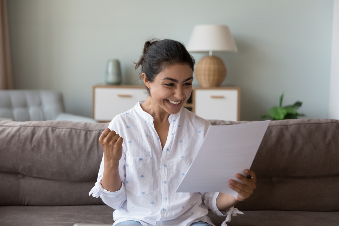 Excited cheerful young Indian woman getting good news letter, reading paper document at home, making winner yes hand, feeling happy, smiling, laughing, celebrating, success, achieve