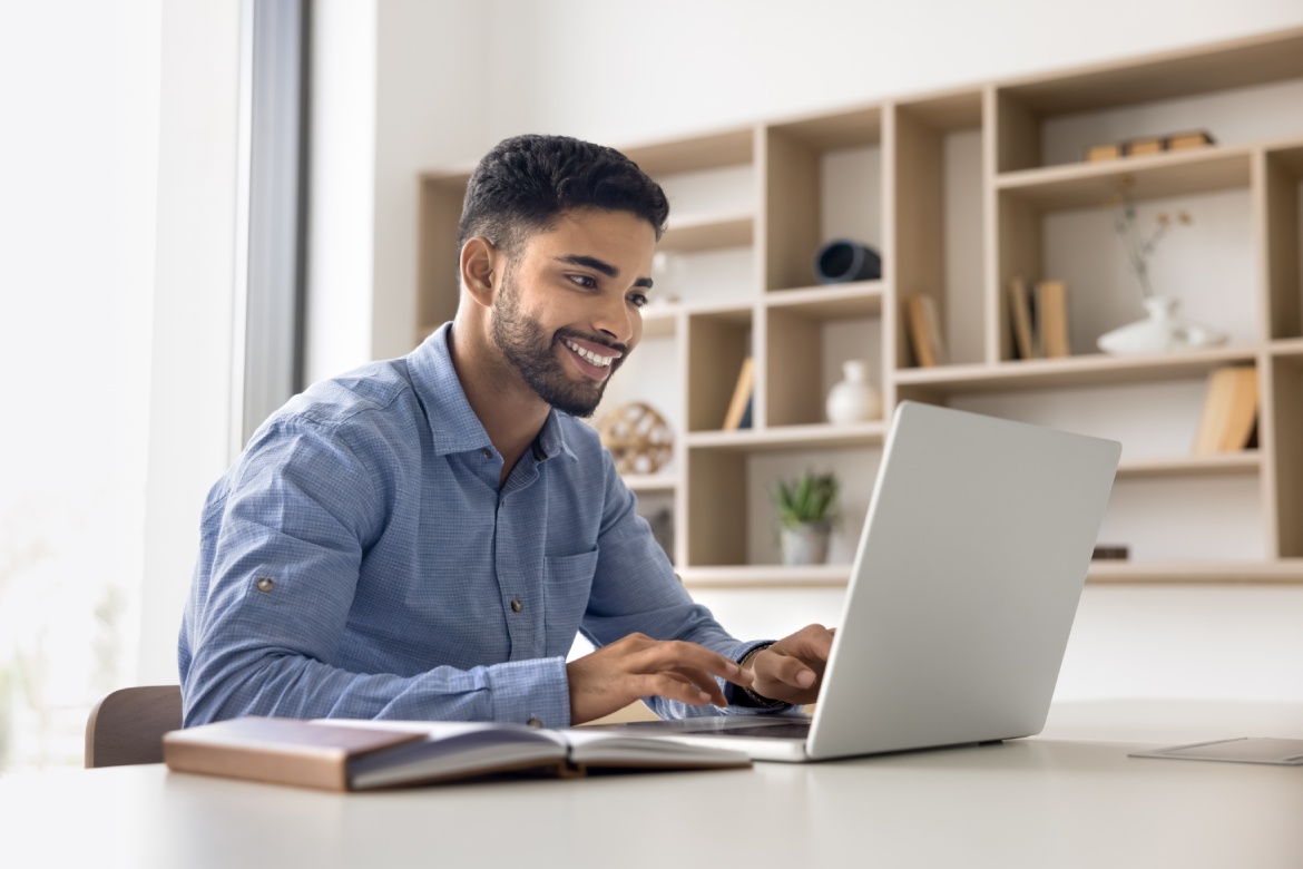 Positive satisfied young Arab employee man working in office alone, sitting at workplace with computer typing on laptop, using online service on pc for job communication, smiling