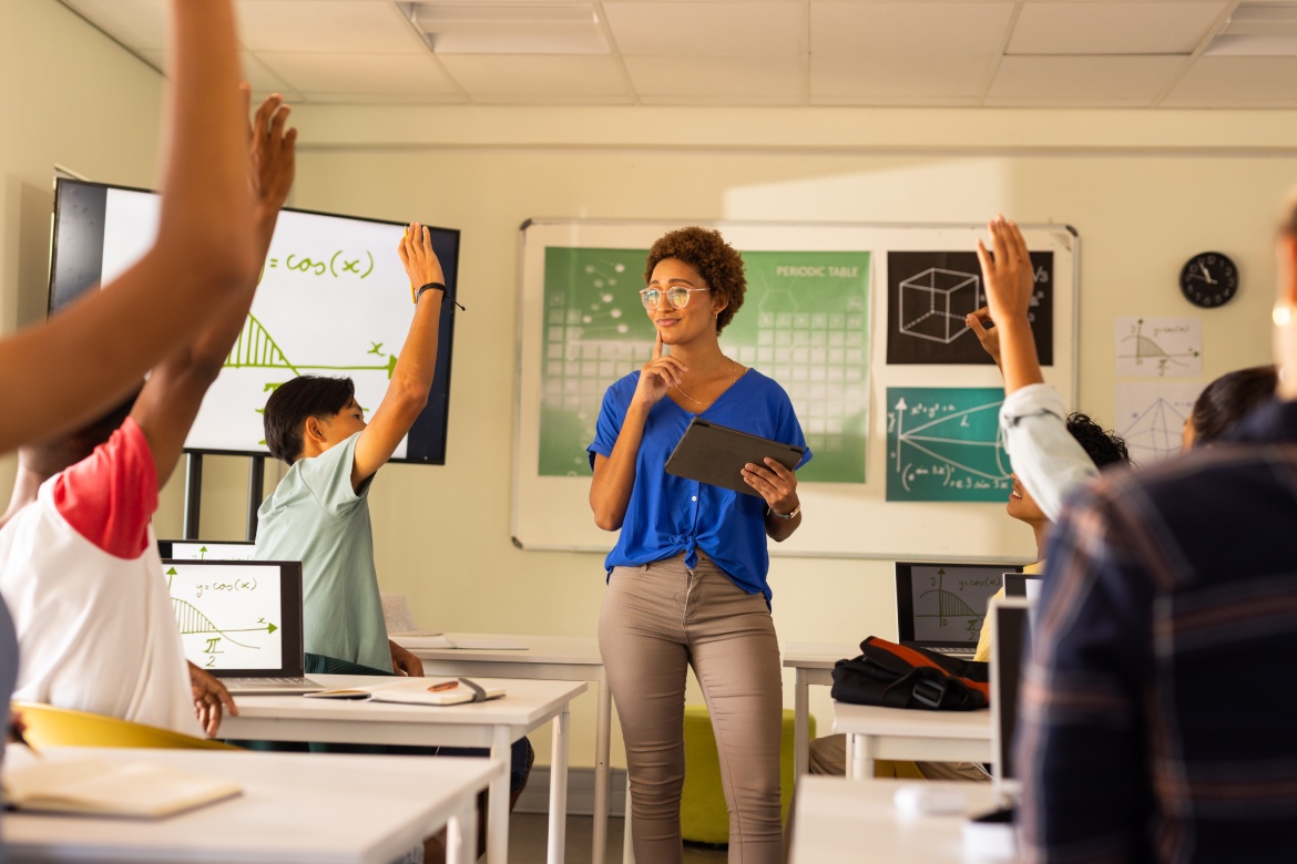 In high school, teacher holding tablet and engaging students raising hands in classroom. Education, teaching, learning, technology, engagement, interaction