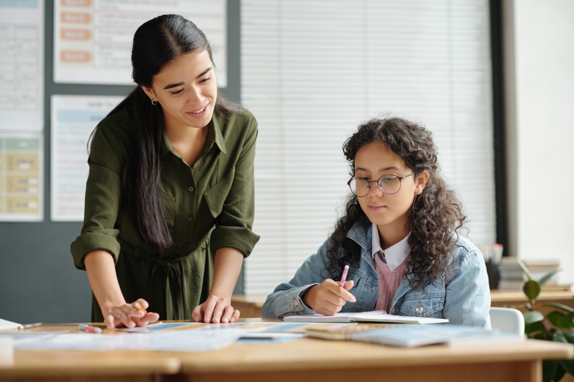 Happy young teacher of English language bending over desk with grammar tables illustrating usage of verb forms and tenses